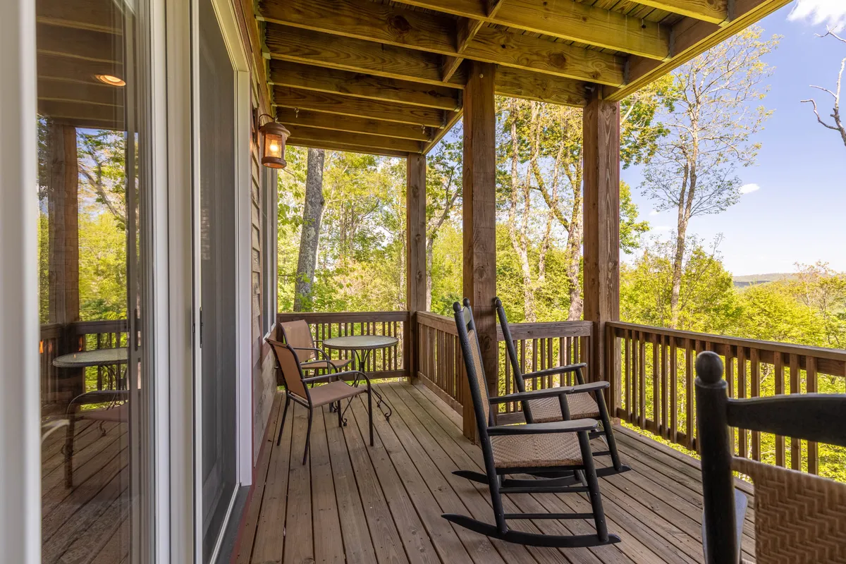 This exterior image features a deck with wooden rocking chairs and a table, surrounded by trees. The setting exudes serenity, offering a tranquil spot for relaxing, reading, or soaking in sunset views and mountain air.