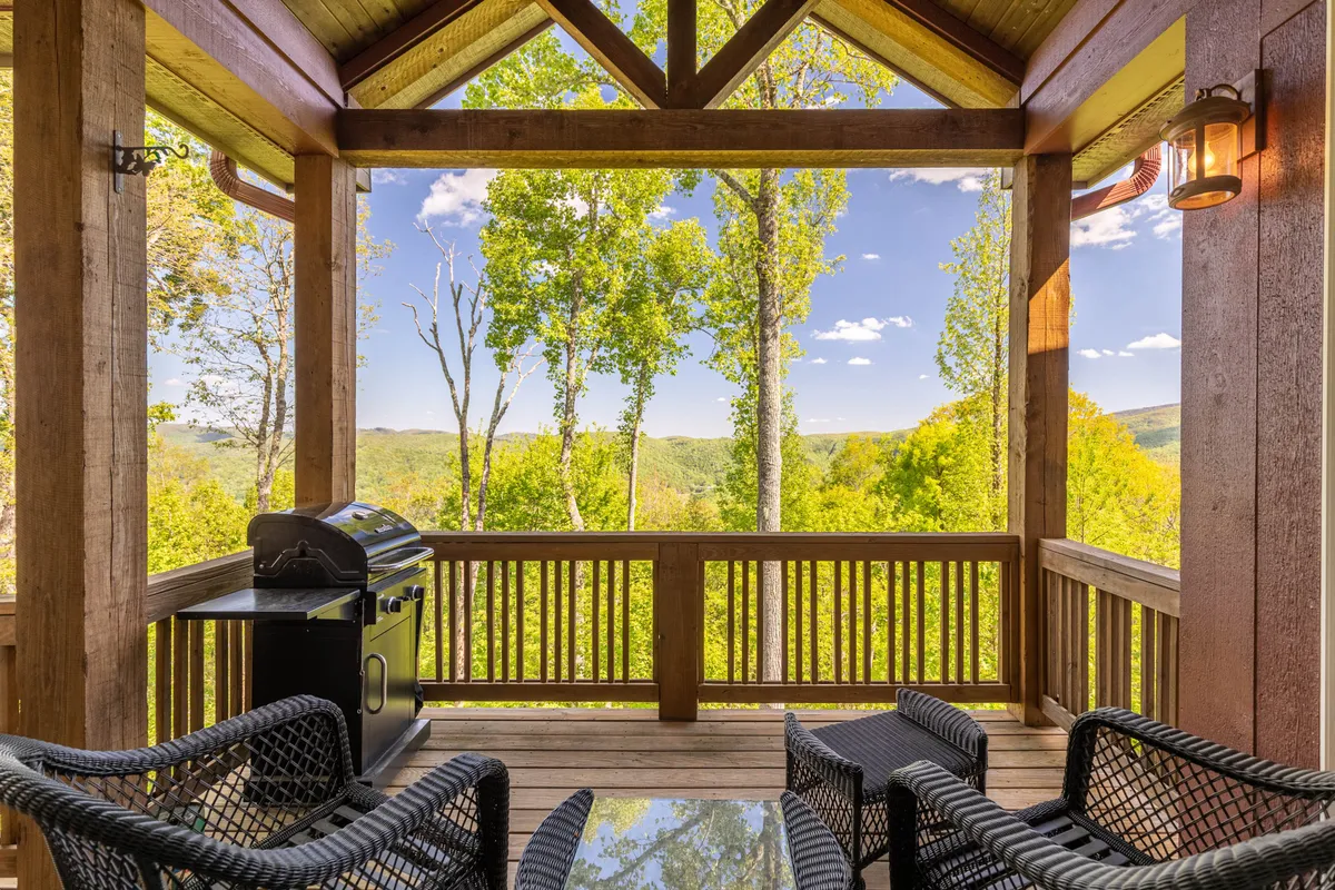 This image shows a spacious deck with a comfortable outdoor seating area, a gas BBQ grill, and a breathtaking panoramic view of trees and mountains. The scene is bathed in warm light, suggesting either morning or sunset, inviting relaxation and outdoor gatherings.