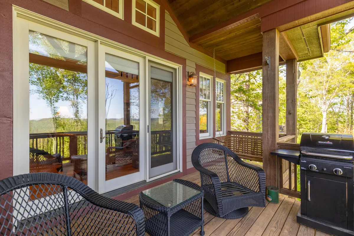 A cozy deck area featuring patio furniture and a gas grill, surrounded by natural greenery and large windows leading back into the house. The setting is relaxed and ideal for enjoying a meal outdoors or morning coffee while immersed in nature.