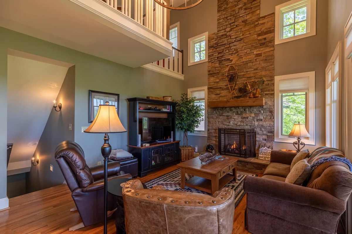 A warm, comfortable living room with a prominent stone fireplace as its centerpiece. Ample natural light pours in through large windows, illuminating the inviting seating area arranged around a coffee table, with a glowing fire enhancing the cozy atmosphere.