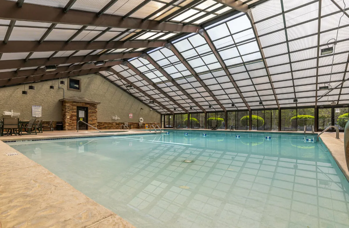 An indoor swimming pool with a large glass ceiling, ample natural light, lounge chairs, and a stone fireplace visible in the background, suggesting a relaxing and luxurious environment for swimming or lounging.