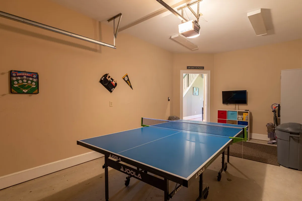 A brightly lit garage space featuring a ping pong table positioned in the center, colorful wall decorations, a mounted TV, and a visible garage door opener. The area is set up for friendly games and relaxed gatherings, with ample space for sporting activities.