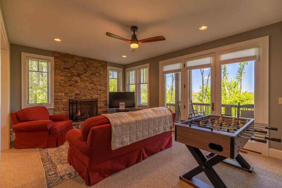 A spacious game room with a stone fireplace, vibrant red armchairs, a foosball table, and large windows letting in natural light. The setting is casual yet inviting, perfect for game nights or relaxing between rounds.