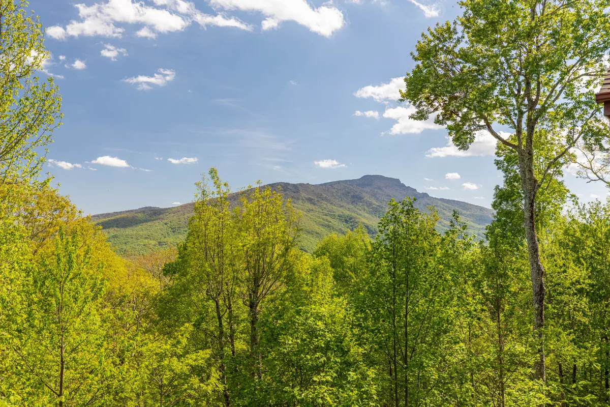 A classic mountain view scene with dramatic peaks, lush green trees, and a clear blue sky. The image captures the sense of openness and tranquility that defines North Carolina’s High Country, as seen from the deck.
